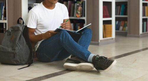 Teenage male student sitting on the floor of a library, writing in a notebook.