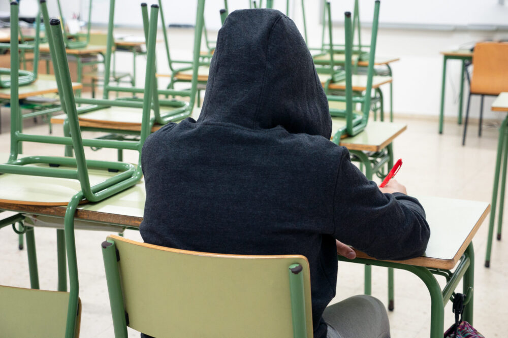Student wearing a black hoodie sits alone in a classroom, back to the camera.