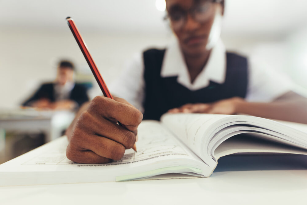 Close-up of a teenage girl writing in her textbook while sitting at desk in classroom. (Photo by Jacob Lund, Adobe Stock)