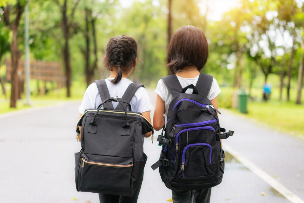 Two young girls with backpacks, viewed from behind, walking to school.