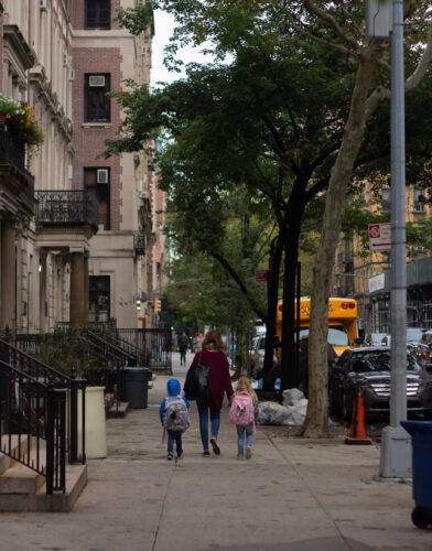 Woman holding the hands of two small children wearing backpacks and walking down a residential NYC street, viewed from behind.