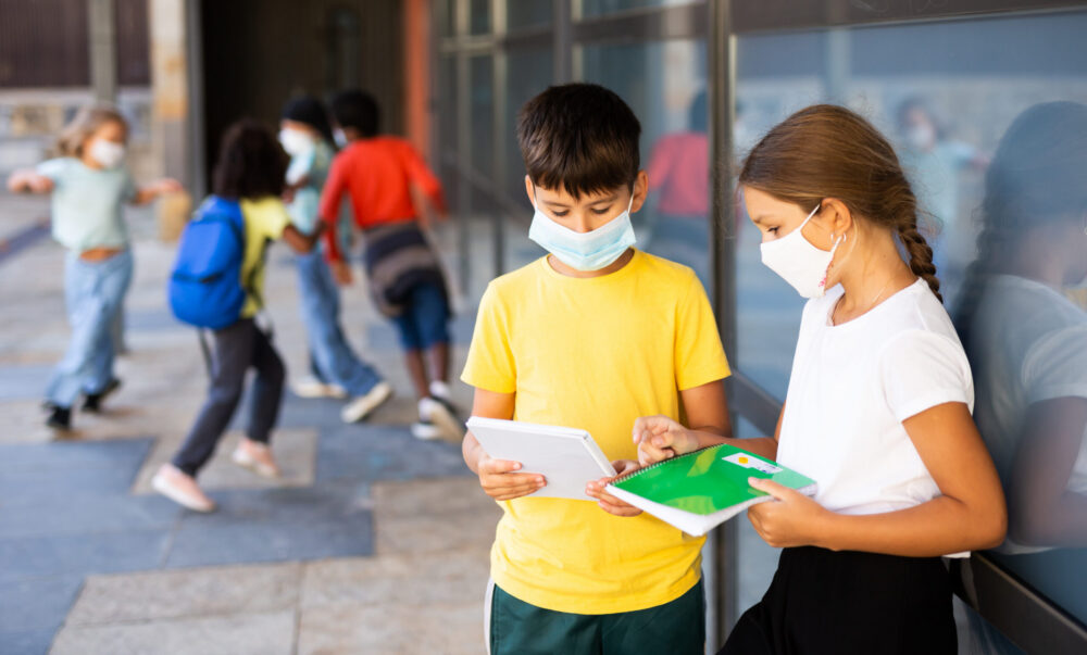 Boy and girl in face masks looking at book talking about homework after classes near school