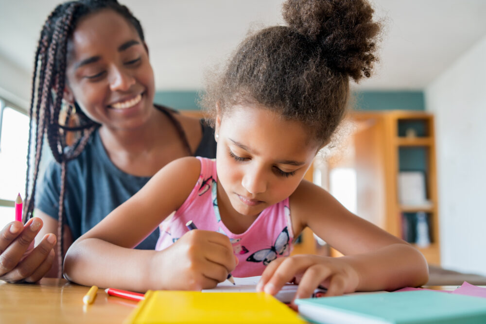 Elementary-school-age child sitting with her mother and writing in a notebook.