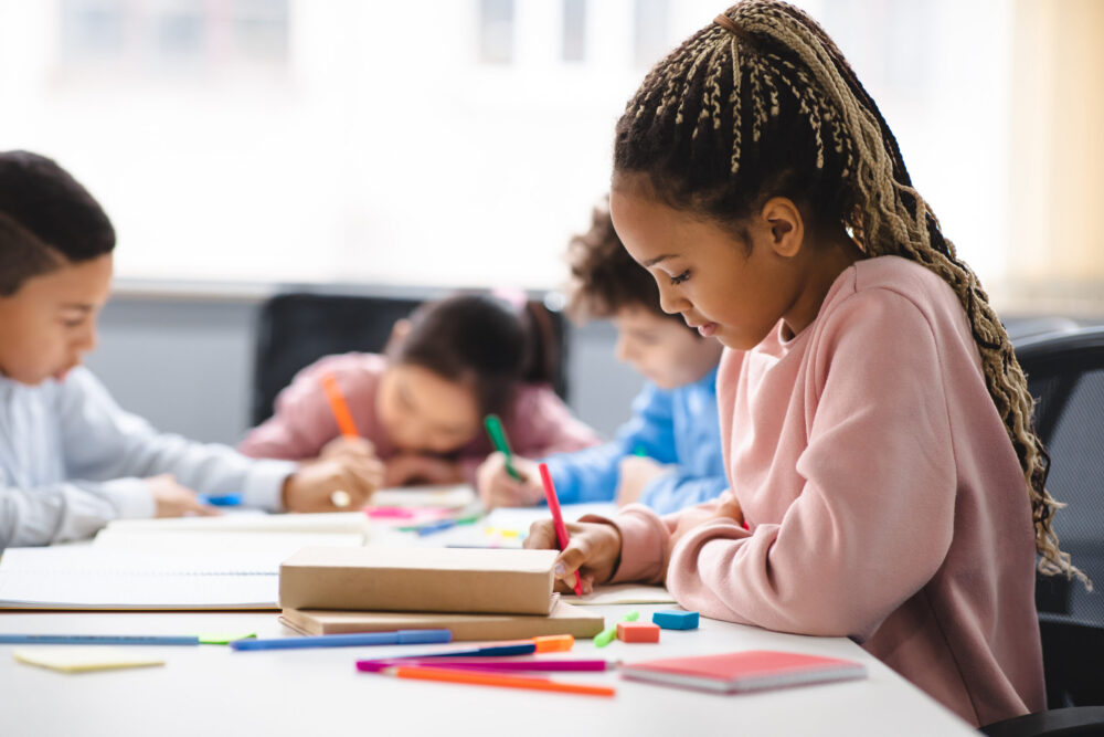 Young Black girl sitting at a table with other students and working on an assignment