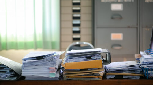Cluttered desk with stacked documents and file cabinets in background