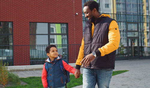 Happy little boy with backpack holding hands with dad, walking home after school