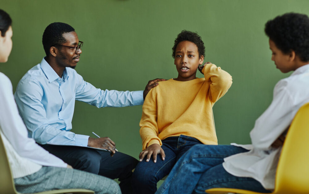 Dissatisfied schoolboy touching back of head while describing his problem at session and school psychologist supporting him.