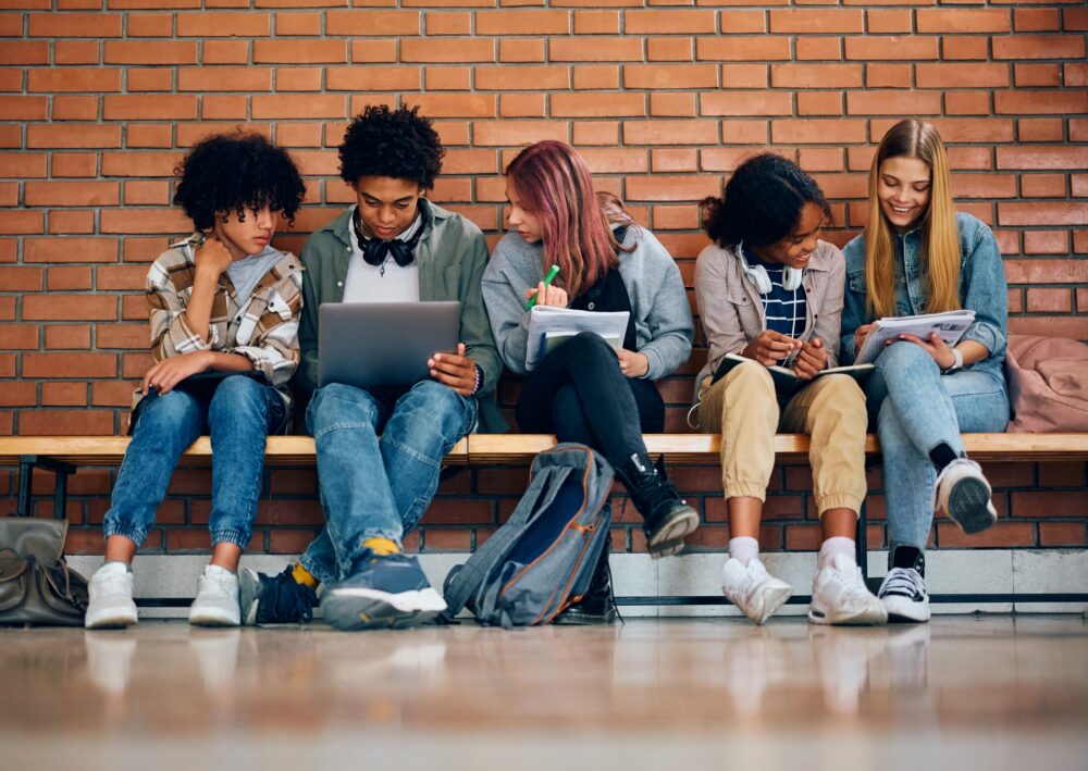 Five teenage students sitting on a bench in front of a brick wall, looking at notebooks and a laptop.
