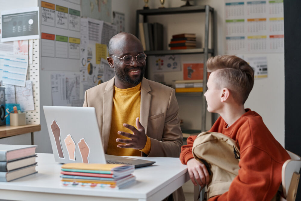 Smiling Black teacher sitting at desk, gesturing at laptop while talking with a student