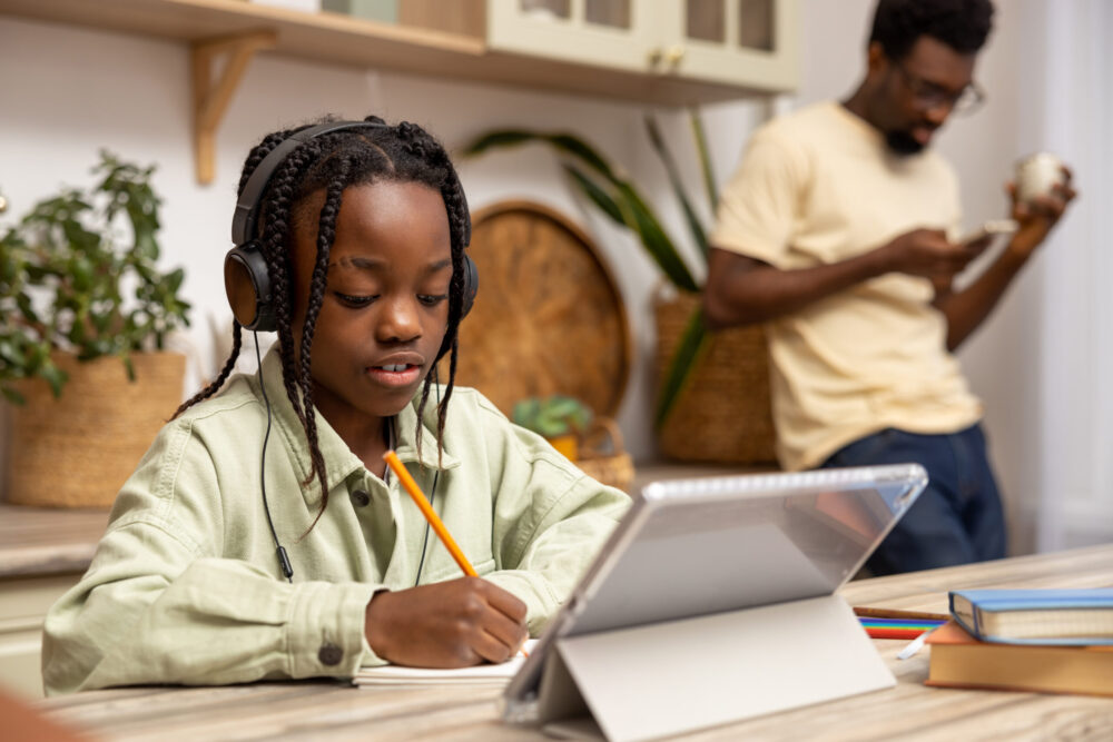 Young Black girl taking class online via a tablet while her dad stands in the background.