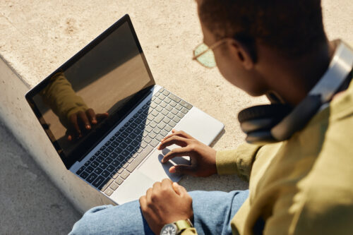 Over shoulder shot of Black teenage boy using laptop on concrete bench in park