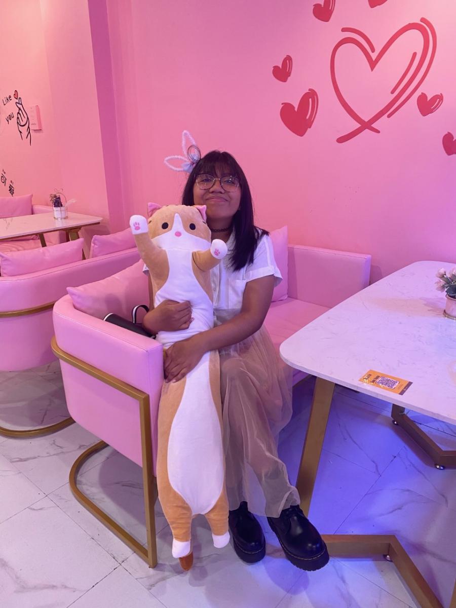 alex sits in a bright pink room holding a kitten stuffed animal, smiling at the camera