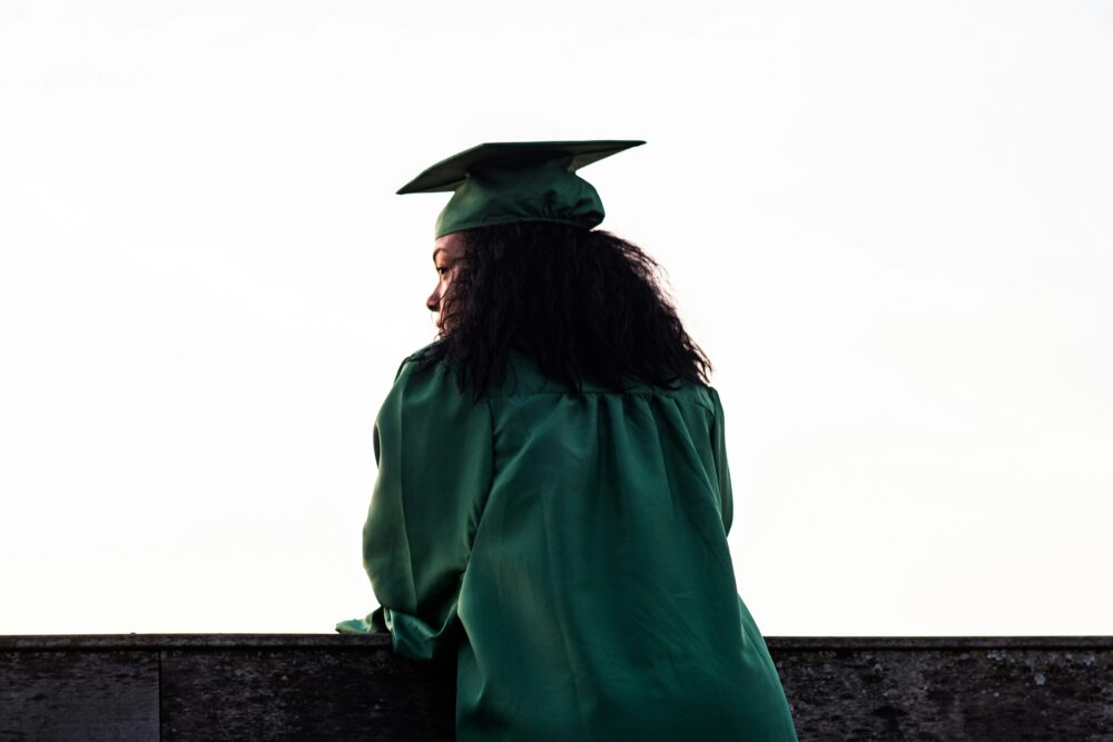Young woman in dark green cap and gown stands with her back to the camera, looking off to the left.