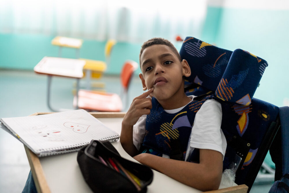 Boy with cerebral palsy in a classroom; an open notebook and pencil case are on his desk.
