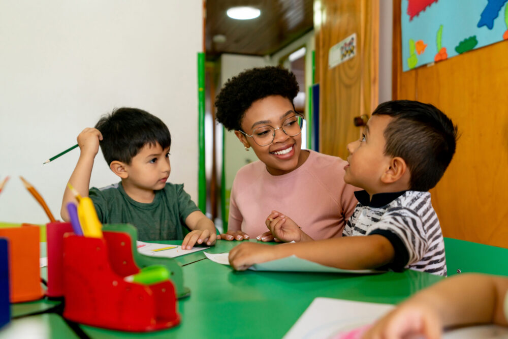 Teacher kneels on the floor between two preschool boys, who are sitting at a table drawing.