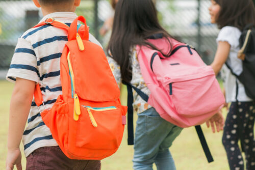 Three young children wearing brightly colored backpacks, viewed from behind.