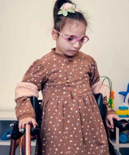Young girl wearing glasses and a brown dress and using a walker.