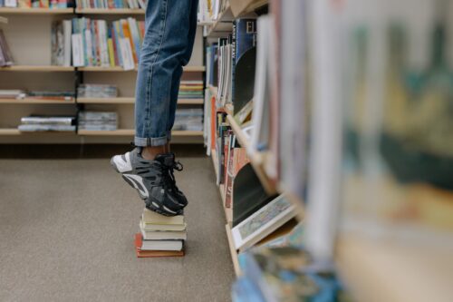 Sneakered feet of a student standing on a pile of books to reach a higher library shelf.
