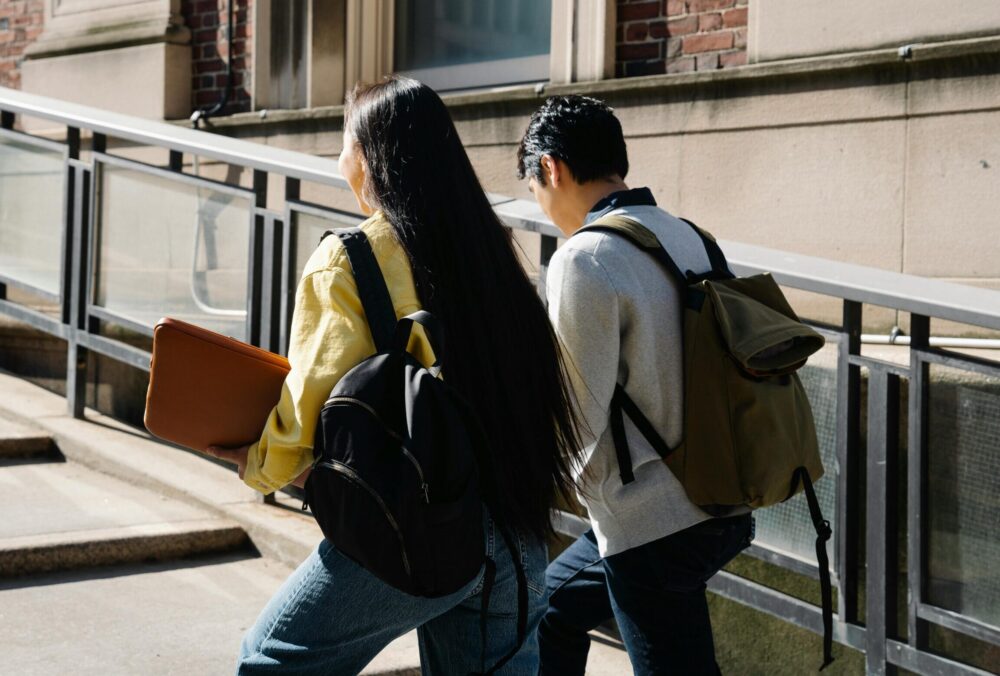 Rear view of two students wearing backpacks walking up an incline. (Photo by George Pak via Pexels)
