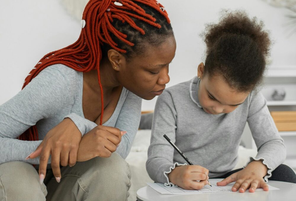 Woman looks over the shoulder of a young girl filling out a worksheet. (Photo by Monstera Production via Pexels)