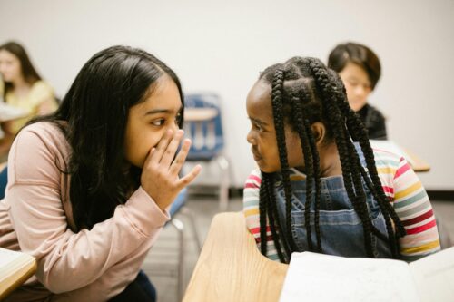 Girl sitting at classroom desk and leaning towards another girl, whispering