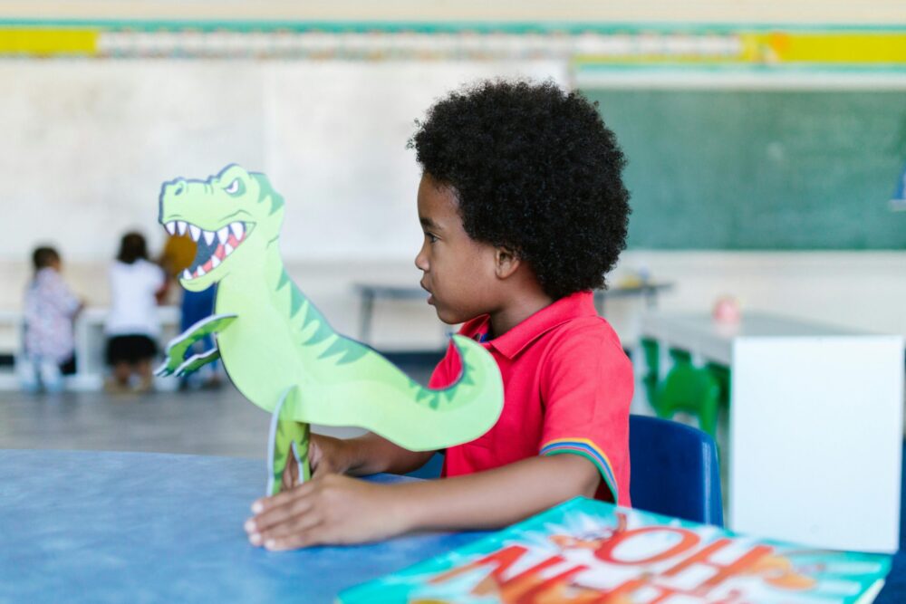 Side view of a Black preschool boy in a classroom, holding a large dinosaur cutout.