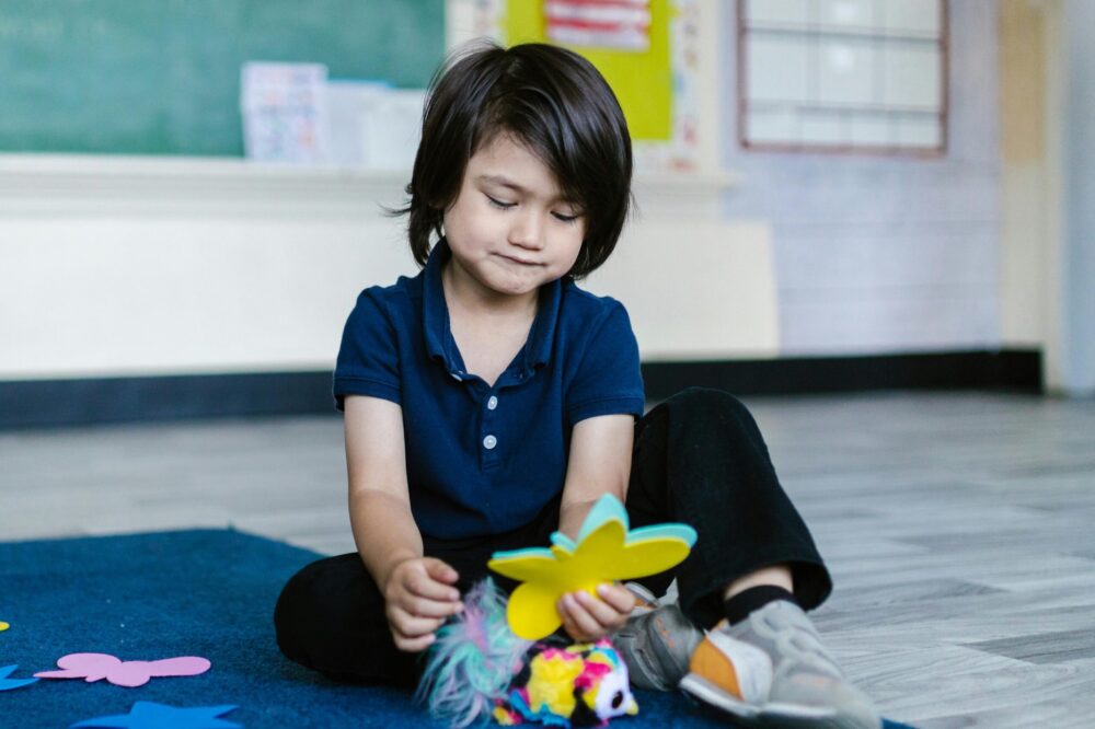 Preschool child sits on the floor of a classroom, looking down at paper cutouts in their hand.