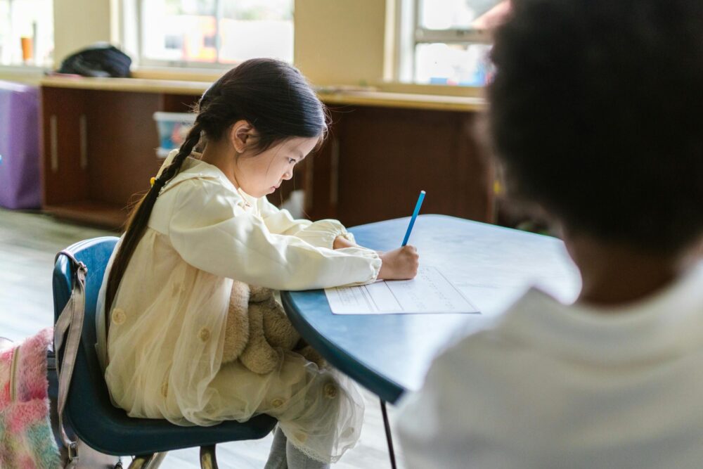 Preschool girl sits at a small table, writing on a piece of paper, a look of concentration on her face.