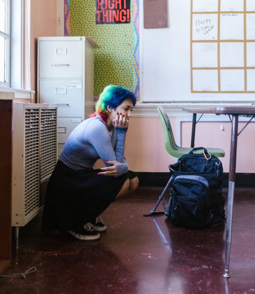 Teen girl with rainbow-colored hair squats in the corner of a classroom, looking at the floor.