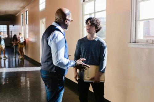 Male teacher speaks with a teenage boy in a school hallway.