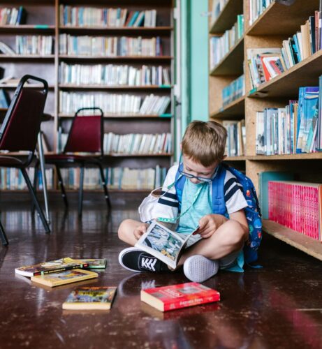 Young boy wearing a backpack sits on the floor of a library, reading a book. A face mask hangs around his neck.