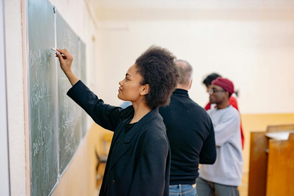 Woman in black jacket writing on chalkboard. (Photo by Yan Krukau via Pexels)
