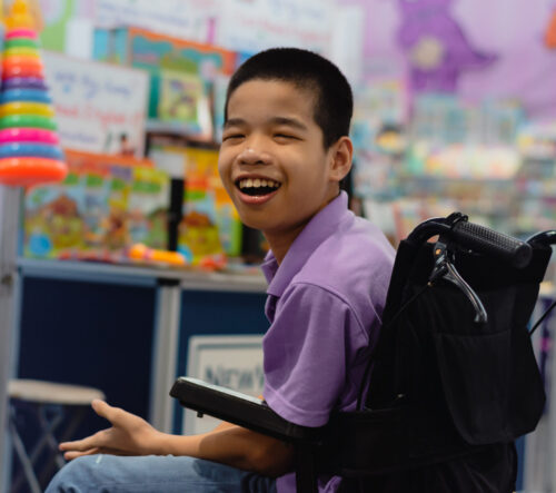 Smiling Asian boy in a wheelchair in a classroom.