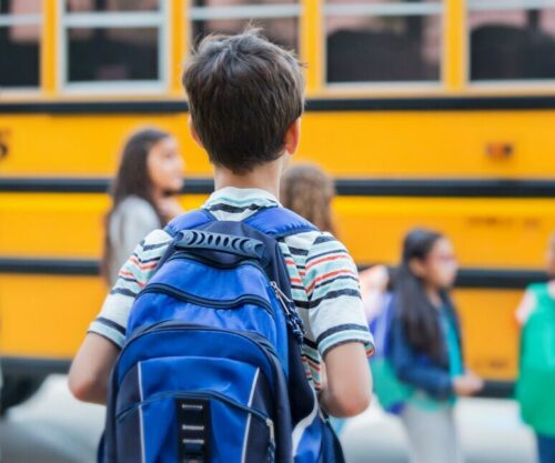 Boy wearing a backpack, viewed from behind walking towards a school bus.