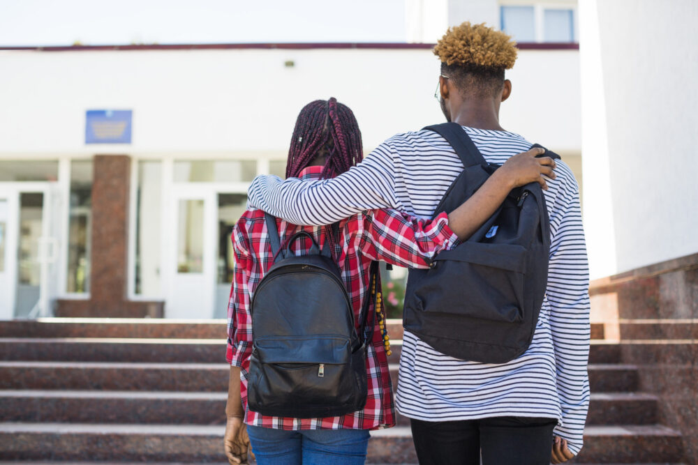 Two teenagers walk up the steps of a school, arms around one another.