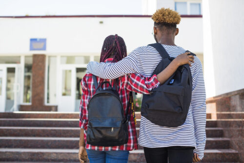 Two teenagers walk up the steps of a school, arms around one another.